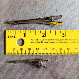 May include: Two silver alligator hair clips, each measuring approximately 5.5 cm in length, are shown against a gray fabric background. A yellow ruler is positioned below the clips for size reference.