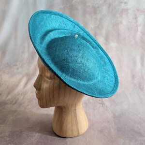 May include: A teal-coloured hat with a circular design, featuring a slightly raised centre and a small, decorative accent. The hat is displayed on a wooden head form, showcasing its shape and texture. The hat's brim is approximately 30 cm in diameter.