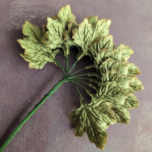 May include: A cluster of artificial leaves in shades of green and brown, arranged on a green wire stem. The leaves have a textured, velvety appearance, and the stem is tightly wrapped. The arrangement is likely for floral arrangements or craft projects.