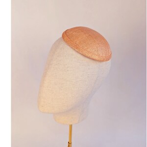 May include: A small, round, peach-colored hat made of woven straw. The hat is sitting on a white mannequin head.