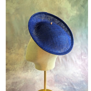 May include: A navy blue, saucer-style hat with a textured weave. The hat is displayed on a mannequin head with a gold-coloured stand. A small, light-coloured embellishment is on the hat's surface. The hat is a formal accessory.
