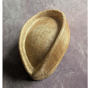 May include: A golden-tan, oval-shaped fascinator hat featuring a structured, woven straw-like texture. The hat has a raised, curved brim that tapers to a point at one end, creating a distinct, asymmetrical silhouette with a smooth, uniform finish.