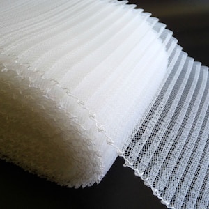 May include: White pleated tulle trim with a scalloped edge. The trim is folded and stacked, showing the texture of the fabric.