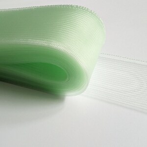 May include: A roll of pale green tulle ribbon with a delicate, textured weave. The ribbon is rolled up and partially unrolled, showing the soft, airy fabric.