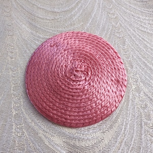 May include: A pink woven straw hat with a circular shape. The hat is made of a tightly woven straw material and has a smooth, even texture.