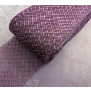 May include: A roll of mauve ribbon featuring a diamond-patterned weave. The ribbon is tightly wound, showcasing the texture and colour variations. The fabric has a delicate, semi-transparent appearance, suitable for crafts or embellishments.