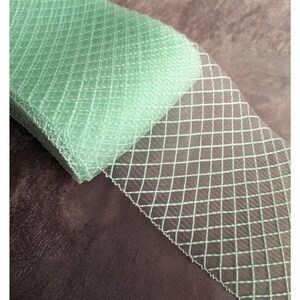 May include: A roll of pale green fabric featuring a diamond grid pattern. The fabric is neatly folded, showcasing the texture and design. This fabric is suitable for crafting, sewing, or decorative applications.