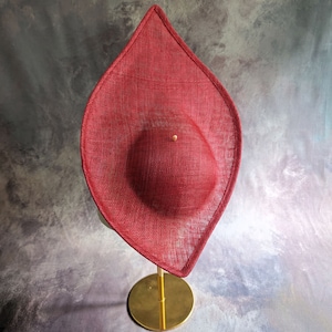 May include: A red, wide-brimmed hat with a rounded crown, displayed on a gold stand. The hat is made of a woven material and has a slightly pointed shape.