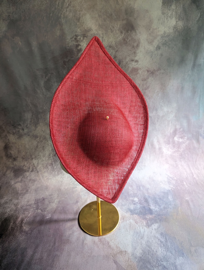 May include: A red, wide-brimmed hat with a rounded crown, displayed on a gold stand. The hat is made of a woven material and has a slightly pointed shape.