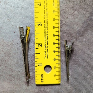 May include: Two silver hair clips, each measuring approximately 4 cm in length, are placed next to a yellow ruler. The ruler is marked in inches and centimeters.