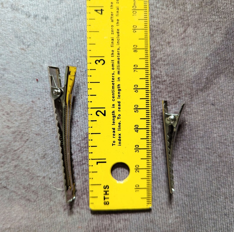May include: Two silver hair clips, each measuring approximately 4 cm in length, are placed next to a yellow ruler. The ruler is marked in inches and centimeters.