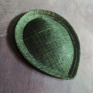 May include: A green, round, hat base made of woven straw. The base is slightly oval shaped and has a smooth, textured surface.