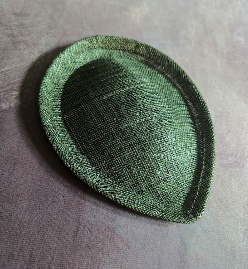 May include: A green, round, hat base made of woven straw. The base is slightly oval shaped and has a smooth, textured surface.