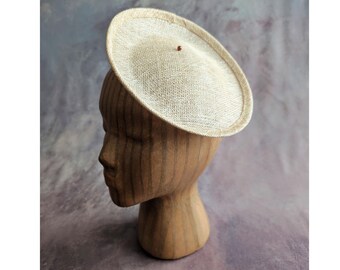 May include: A beige, circular hat with a small, dark brown accent. The hat is made of a woven material and is displayed on a wooden head form. The hat's brim is slightly curved, and the overall design is simple and elegant.