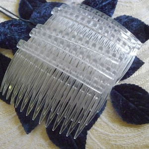 May include: A stack of clear plastic hair combs with long, thin teeth. The combs are arranged on a background of dark blue velvet leaves and a cream-colored floral patterned fabric.