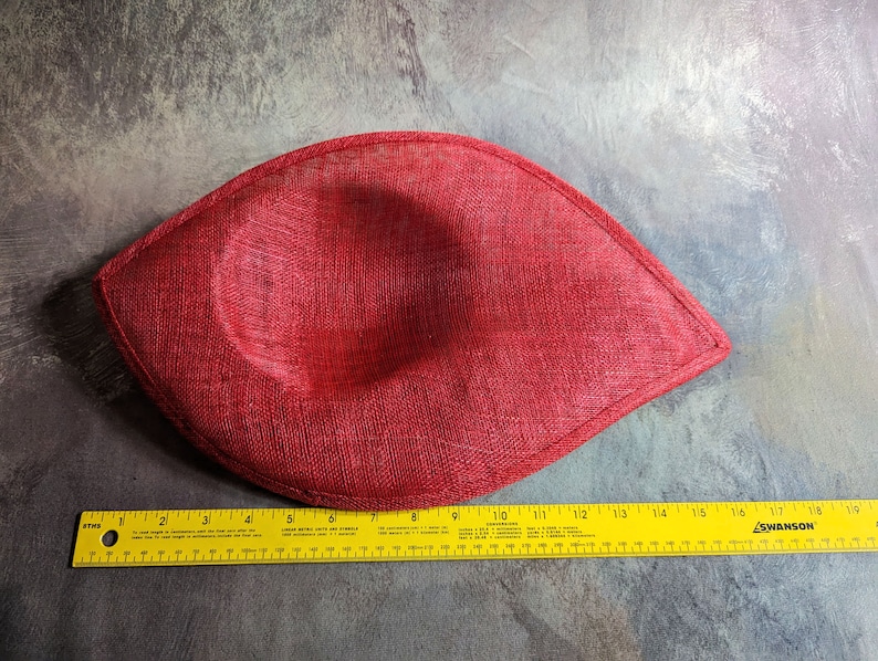 May include: A red fabric hat with a rounded shape and a slightly pointed tip. The hat is measured with a ruler, showing it is about 18 inches long.