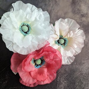 K&ouml;nnte beinhalten: Drei k&uuml;nstliche Blumen, eine wei&szlig;e, eine rosa und eine rote, mit gr&uuml;nen Zentren. Die Blumen sind aus Papier oder Stoff und haben eine zarte, gekr&auml;uselte Textur.