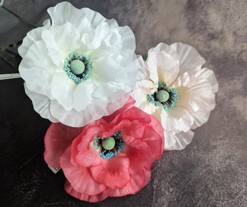 K&ouml;nnte beinhalten: Drei k&uuml;nstliche Blumen, eine wei&szlig;e, eine rosa und eine rote, mit gr&uuml;nen Zentren. Die Blumen sind aus Papier oder Stoff und haben eine zarte, gekr&auml;uselte Textur.
