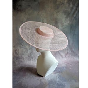 May include: A light pink, wide-brimmed hat with a circular crown and a large, open weave brim. The hat is displayed on a white mannequin head against a muted, textured background. The hat's design suggests it is suitable for formal events.