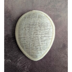 May include: A cream-colored, oval-shaped hat base with a woven texture. The hat base is trimmed with a matching cream-colored border. This millinery supply is suitable for creating custom hats and fascinators.