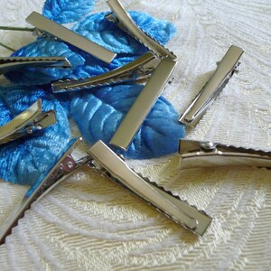 May include: A group of silver alligator hair clips with a scalloped edge. The clips are scattered on a white fabric background.