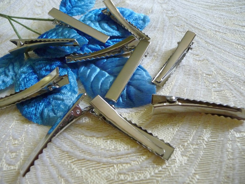 May include: A group of silver alligator hair clips with a scalloped edge. The clips are scattered on a white fabric background.
