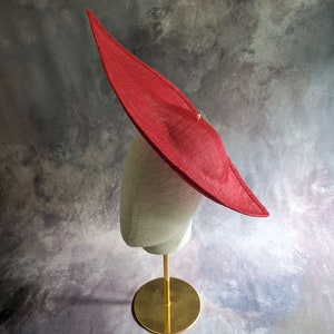 May include: A large, red, wide-brimmed hat sits on a mannequin head. The hat is made of a woven material and has a slightly pointed crown. The mannequin is on a gold stand.