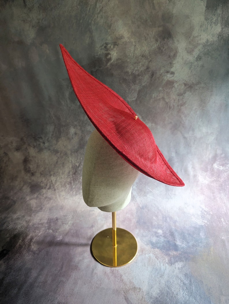 May include: A large, red, wide-brimmed hat sits on a mannequin head. The hat is made of a woven material and has a slightly pointed crown. The mannequin is on a gold stand.