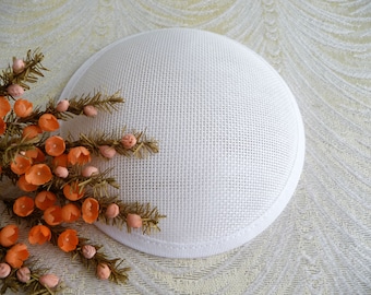 May include: White round hat base made of a woven fabric with a white satin trim. The base is sitting on a white textured surface.