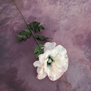 K&ouml;nnte beinhalten: Eine einzelne wei&szlig;e Mohnblume mit einem t&uuml;rkisfarbenen Zentrum auf einem violetten Hintergrund.