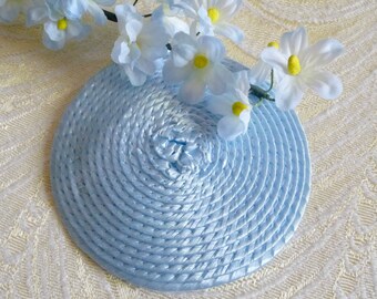 May include: A light blue, round, woven hat with a circular design. The hat is decorated with a cluster of white flowers with yellow centers.