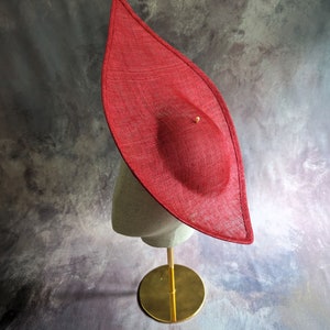 May include: A large, red, wide-brimmed hat made of straw sits on a gold stand. The hat has a pointed crown and a wide, flat brim.