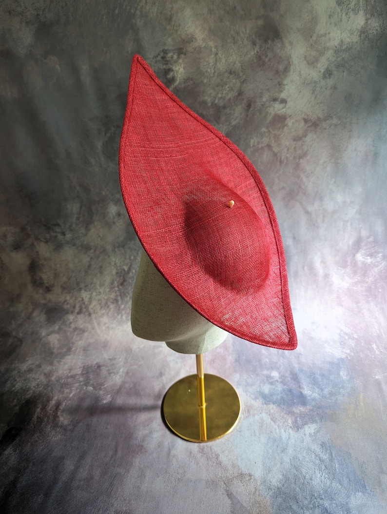 May include: A large, red, wide-brimmed hat made of straw sits on a gold stand. The hat has a pointed crown and a wide, flat brim.