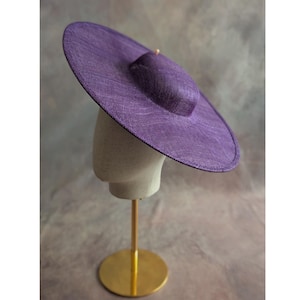 May include: A large, purple sun hat with a wide brim and a rounded crown. The hat is displayed on a mannequin head atop a gold-colored stand. The hat's woven texture is visible, and the overall design is elegant.