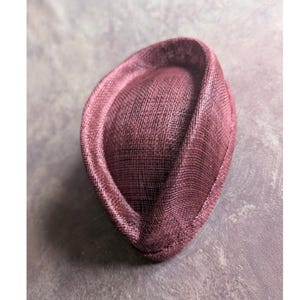 May include: A deep burgundy fascinator hat with a structured, teardrop-shaped crown and a curved, upturned brim. The headpiece features a fine, woven mesh texture throughout, creating a consistent pattern across the entire surface of the hat.