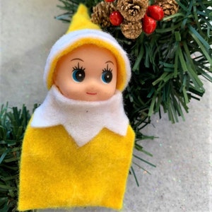 May include: A small doll with a yellow felt outfit and a white and yellow hat. The doll has blue eyes and a small red mouth. The doll is part of a Christmas decoration, with a green garland and pinecones in the background.