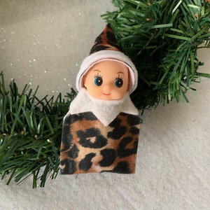 May include: A small elf doll wearing a white hat and a brown and black leopard print outfit. The elf has blue eyes and a smiling face.
