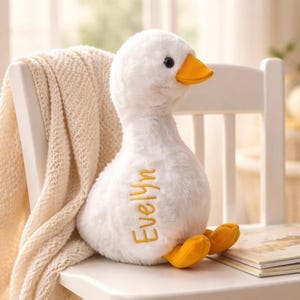 May include: A plush white goose with a yellow beak and feet sits on a white chair. The name "Evelyn" is embroidered in gold thread on the goose's chest. A cream-colored blanket drapes over the chair's back.