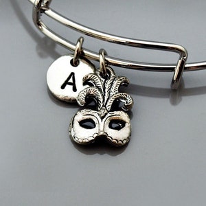 Könnte beinhalten: Ein silbernes Armband mit einem silbernen Buchstaben 'A' Anhänger und einem silbernen Maskenanhänger.