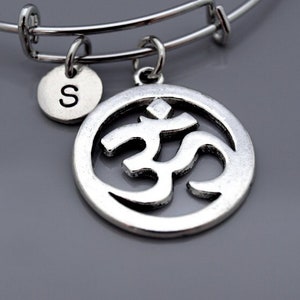 Puede incluir: Un brazalete de plata con un dije de plata con el símbolo Om y un dije de plata con la letra S. El brazalete es expandible.