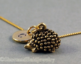 Könnte beinhalten: Goldkette mit einem kleinen Igel-Anhänger und einem runden Anhänger mit dem Buchstaben C. Die Kette liegt auf einer grauen Oberfläche.