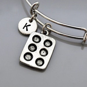 May include: Silver charm bracelet with a letter K charm and a cupcake pan charm. The bracelet is expandable and has a silver finish.