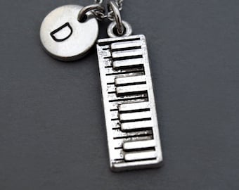 Puede incluir: Collar de plata con un dije de teclas de piano y un dije redondo con la letra D grabada.