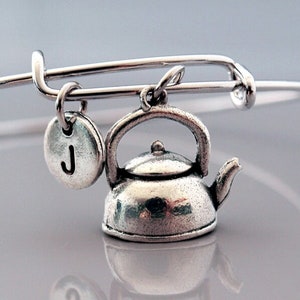 May include: A silver charm bracelet with a teapot charm and a silver charm with the letter J. The bracelet is on a white background.