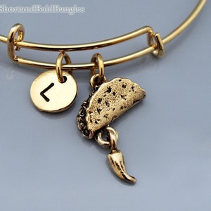 Pode incluir: Um bracelete expansível de ouro com dois pingentes. Um pingente é uma letra inicial "L" de ouro e o outro é um pingente de ouro de um taco com uma pimenta pendurada.