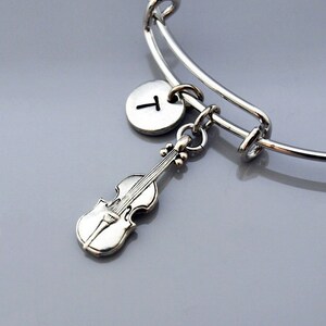 Puede incluir: Un brazalete de plata con un dije de violín de plata y un dije de plata con la letra T.