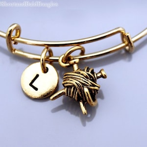 Könnte beinhalten: Goldarmband mit einem Garnknäuel- und Stricknadel-Anhänger und einem runden Anhänger mit dem Buchstaben L.