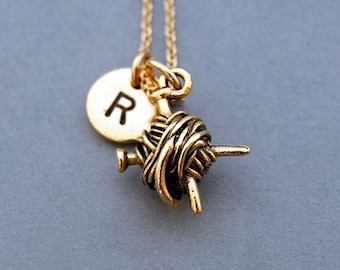 Puede incluir: Collar de oro con un dije de ovillo de lana y aguja de tejer, y un dije redondo con la letra R.