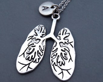 May include: A silver necklace with a charm in the shape of lungs and a small silver disc with the letter A. The necklace is on a gray background.