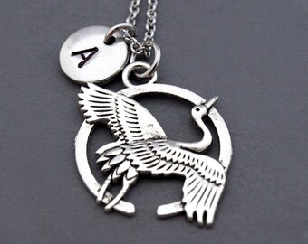 May include: Silver necklace with a crane charm and a small round charm with the letter A. The crane is in flight and the charm is attached to the chain.
